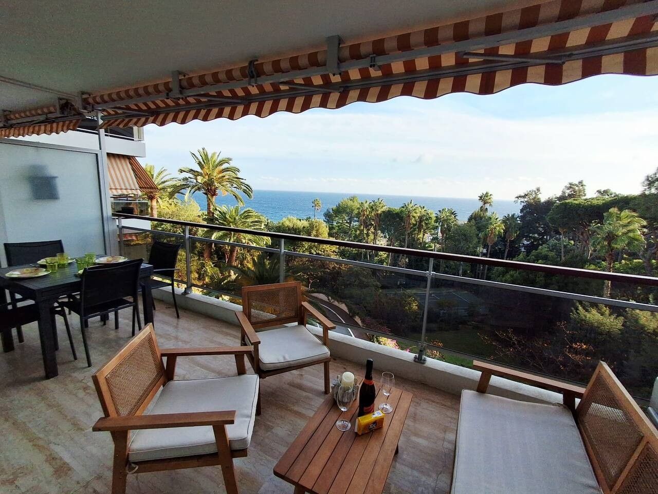 A balcony with wooden furniture, a table with a bottle and glasses, and a view of the ocean and greenery.