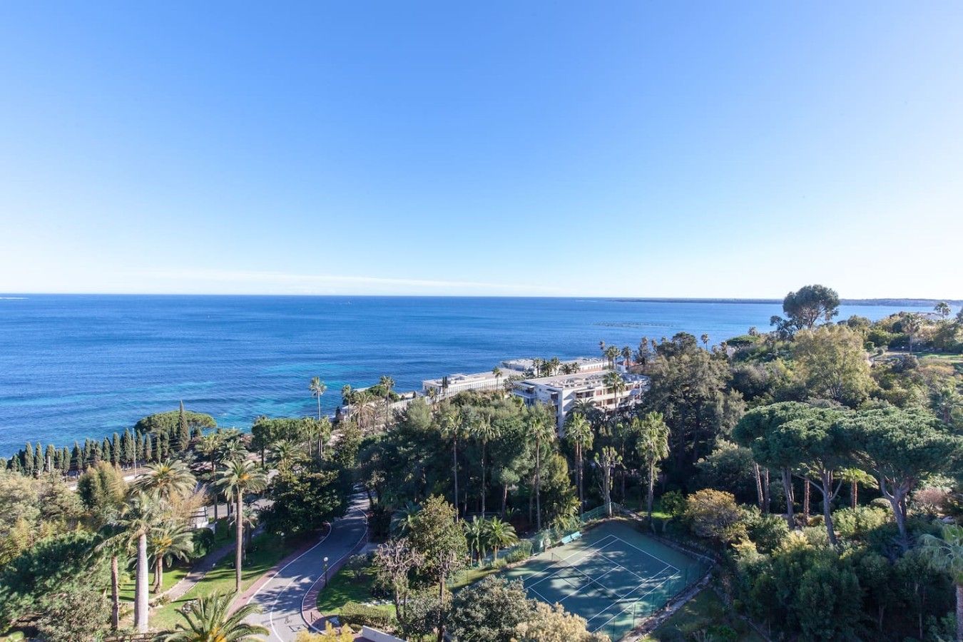 A coastal property with a tennis court, lush greenery, and a view of the sea.