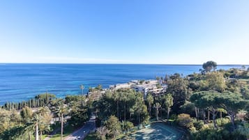 A coastal property with a tennis court, lush greenery, and a view of the sea.