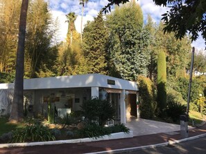 Un piccolo edificio a un piano con facciata bianca, un portico e un'area giardino con diverse piante e un cactus.
