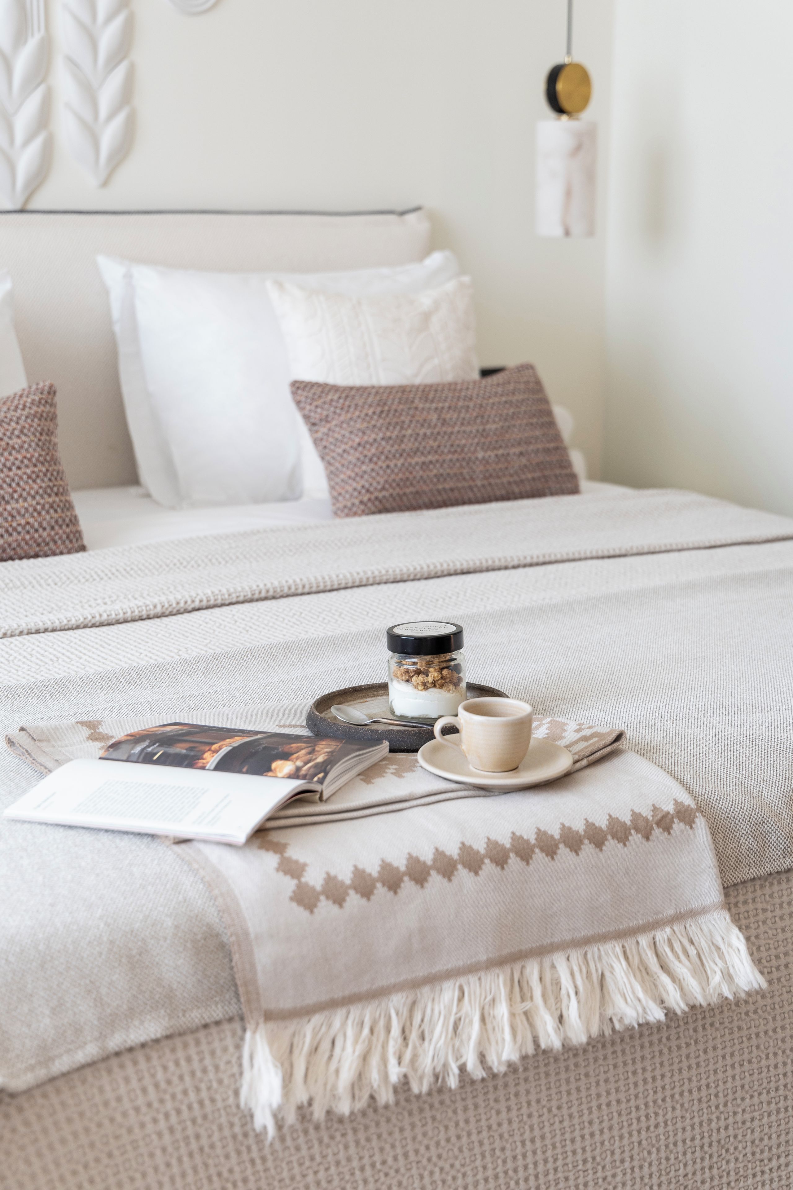 Un letto rifatto con cura, coperto da una coperta bianca e marrone fantasia, un tavolino con una tazza e un vasetto, e una rivista sul letto.