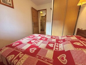 Quarto com cama coberta por um edredom estampado, porta de madeira e corredor que leva a outro cômodo.