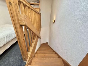 Escada de madeira com corrimão que leva a um quarto com cama e luminária na parede.