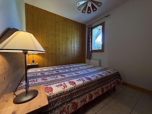 A bedroom with a wooden headboard, a patterned bedspread, a bedside lamp, and a window with curtains. - Ground floor, balcony, tv, ski locker, 35m², Praz sur Arly (Praz-sur-Arly)