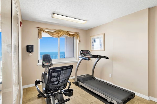 A fitness room with a treadmill, exercise bike, and a window with a view. - Beachfront corner condo w direct gulf access, amazing views and ideal location (Gulf Shores)