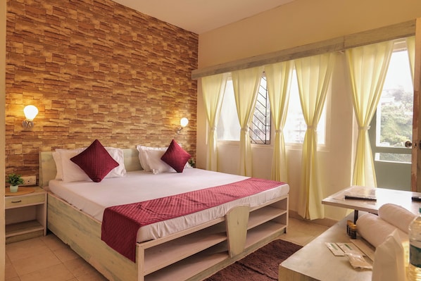 A hotel room with a stone wall, a bed with white bedding and red pillows, a bedside table with a lamp, and a window with curtains.