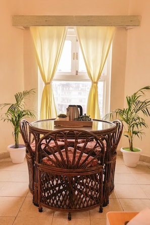 Een ronde rieten tafel met een glazen blad, met aan weerszijden twee potplanten, in een kamer met een raam en gele gordijnen.