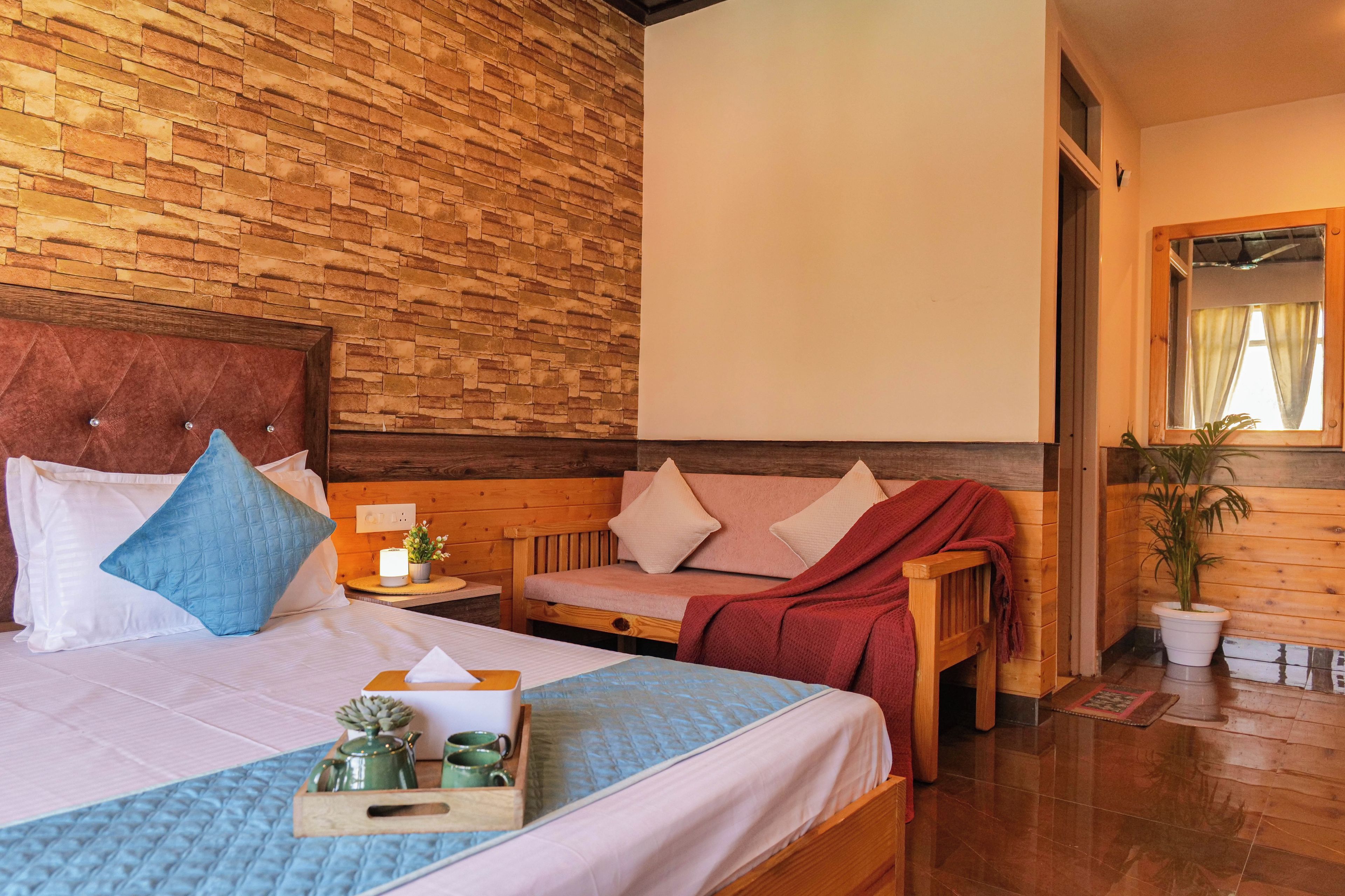 Quarto de hotel com parede de tijolos, cama com travesseiros, mesa de cabeceira com abajur, um banco de madeira e um vaso com planta.