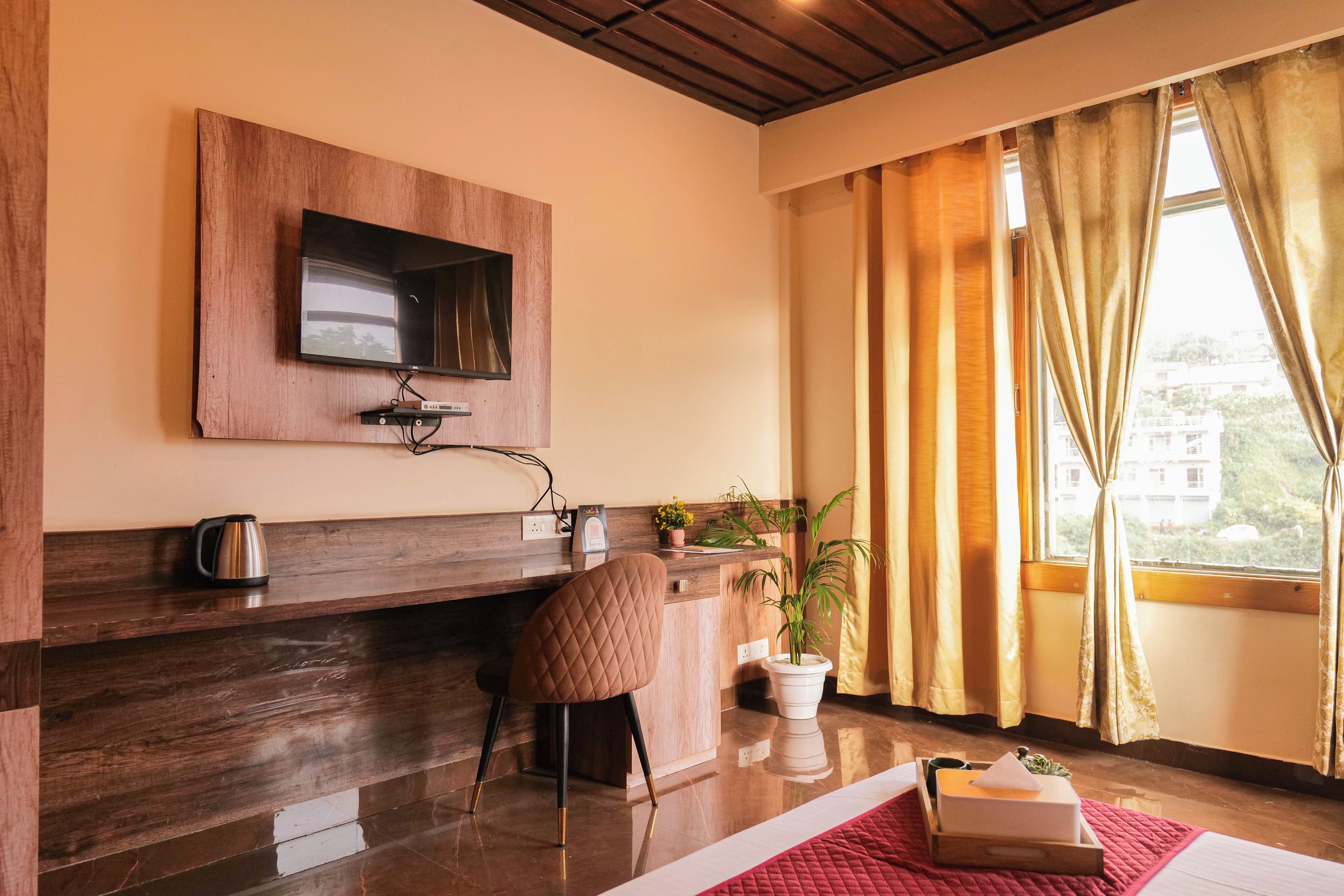Een hotelkamer met een houten bureau, een stoel, een aan de muur bevestigde televisie, een raam met gordijnen en uitzicht op het omliggende landschap.