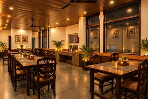A dining area with wooden tables and chairs, potted plants, and artwork on the walls.