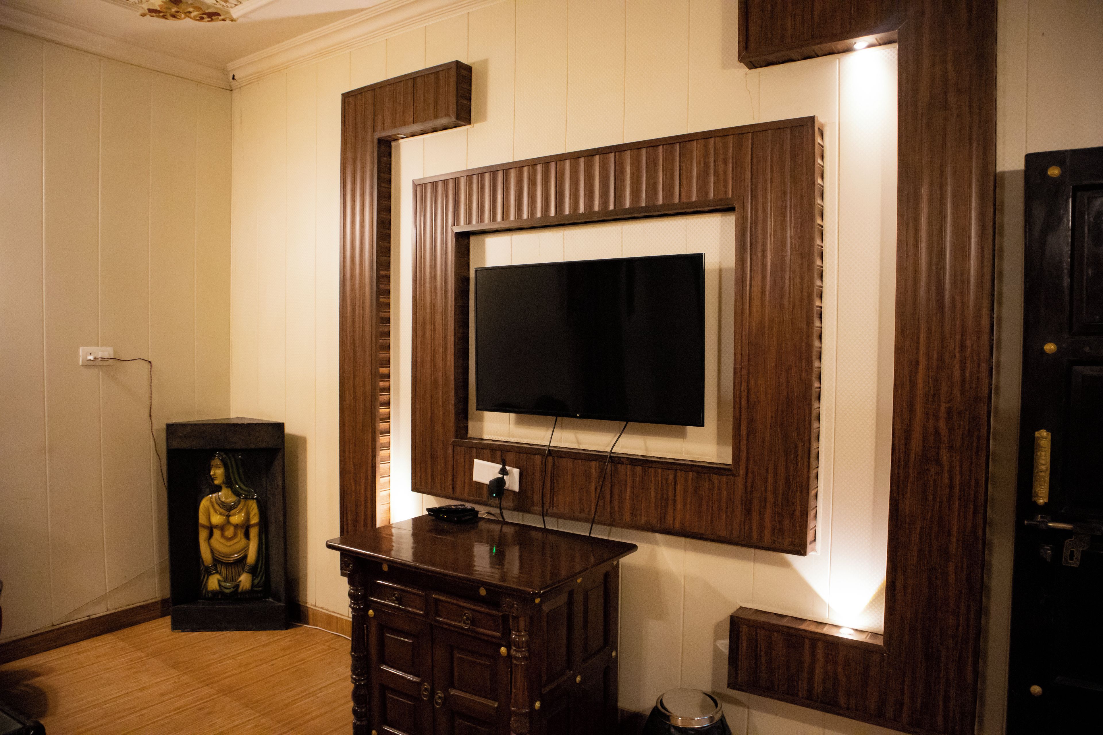 A room with a television mounted on a wooden panel wall, a small statue on a stand, and a door with a handle.