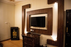 Una habitación con un televisor fijado a una pared de paneles de madera, una pequeña estatua sobre un pedestal y una puerta con manija.