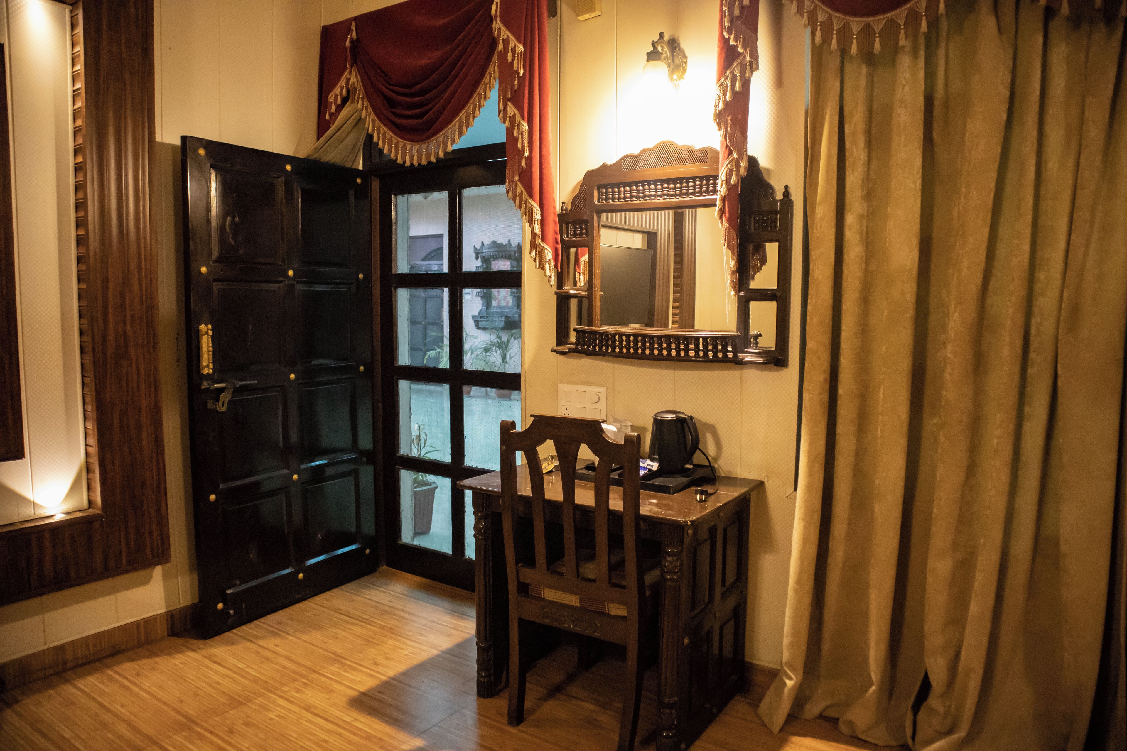 Une pièce avec une porte en bois, une petite table avec une chaise, un miroir fixé au mur et une fenêtre avec des rideaux.