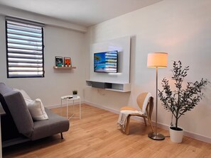 A modern living room with a grey sofa, a small round table with a plant, a floor lamp, and a TV mounted on the wall. - Stay Together! Two Apartments in San Germán! (San Germán)