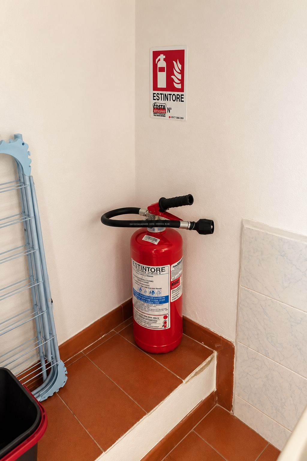 A fire extinguisher mounted on a wall in a corner, with a blue metal ladder leaning against the wall and a red bucket on the floor.