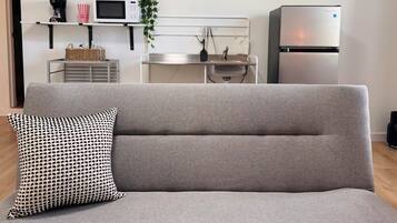 Un salon moderne avec un canapé gris, un coussin à motifs, un coin cuisine équipé d’un four à micro-ondes et d’un réfrigérateur, ainsi qu’une petite table avec un panier.