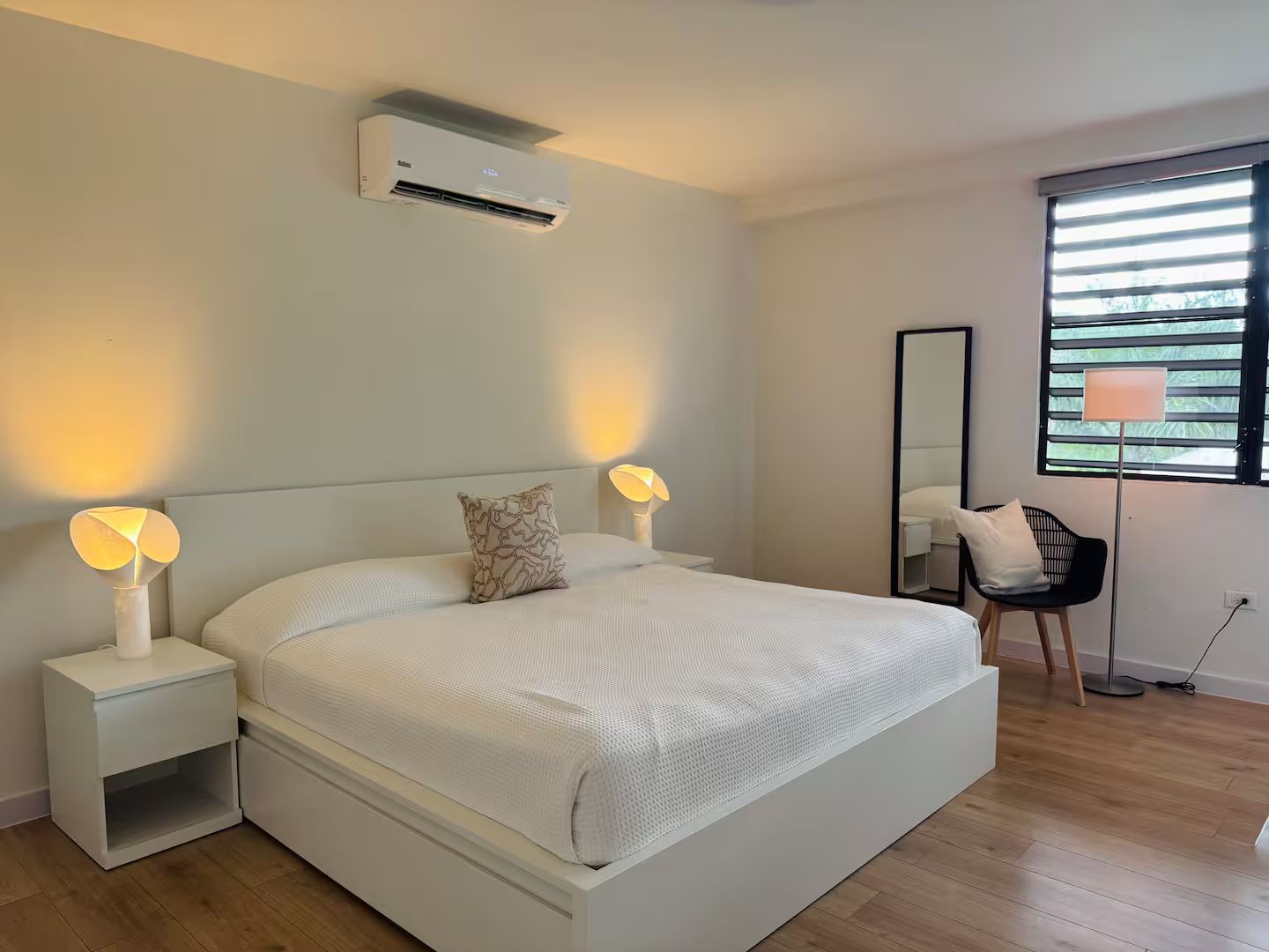 Un dormitorio moderno con una cama grande, lámparas de noche, una lámpara de pie, una silla y una ventana con persianas.