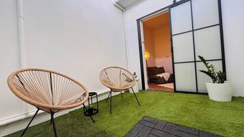 Un pequeño espacio exterior con césped artificial, dos sillas de mimbre, una planta en maceta y un puertas corredera que da a un ambiente interior.