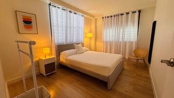 A hotel room with a bed, two bedside tables, a chair, a floor lamp, and a wall-mounted air conditioning unit.