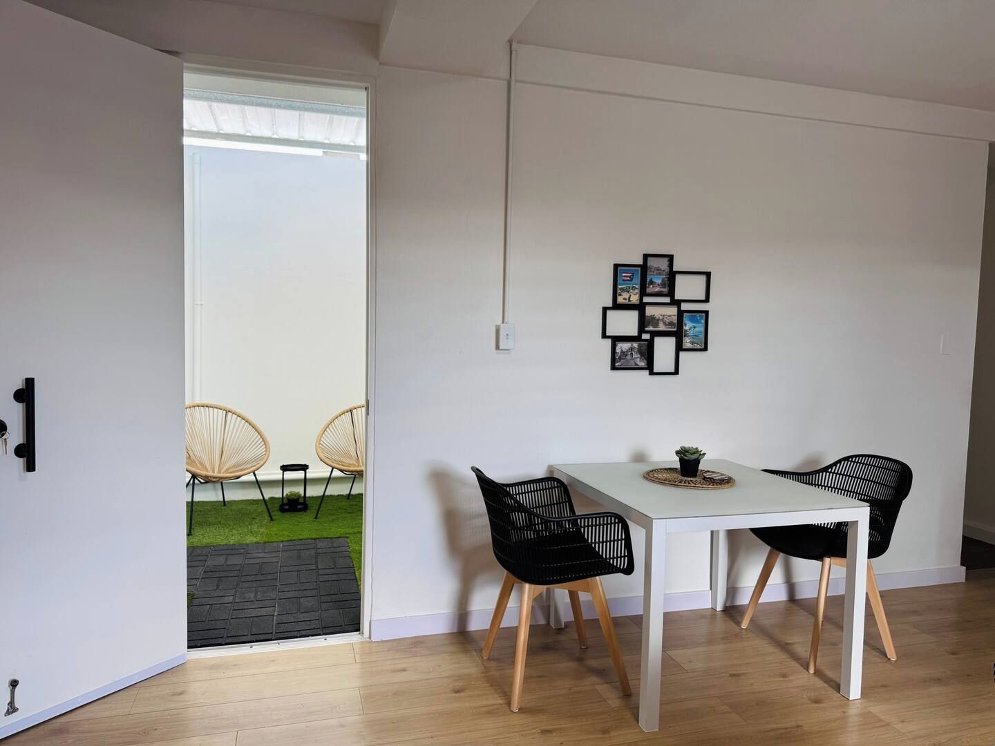 Un comedor moderno con una mesa blanca y sillas negras, una pequeña planta en maceta y un cuadro colgado en la pared.