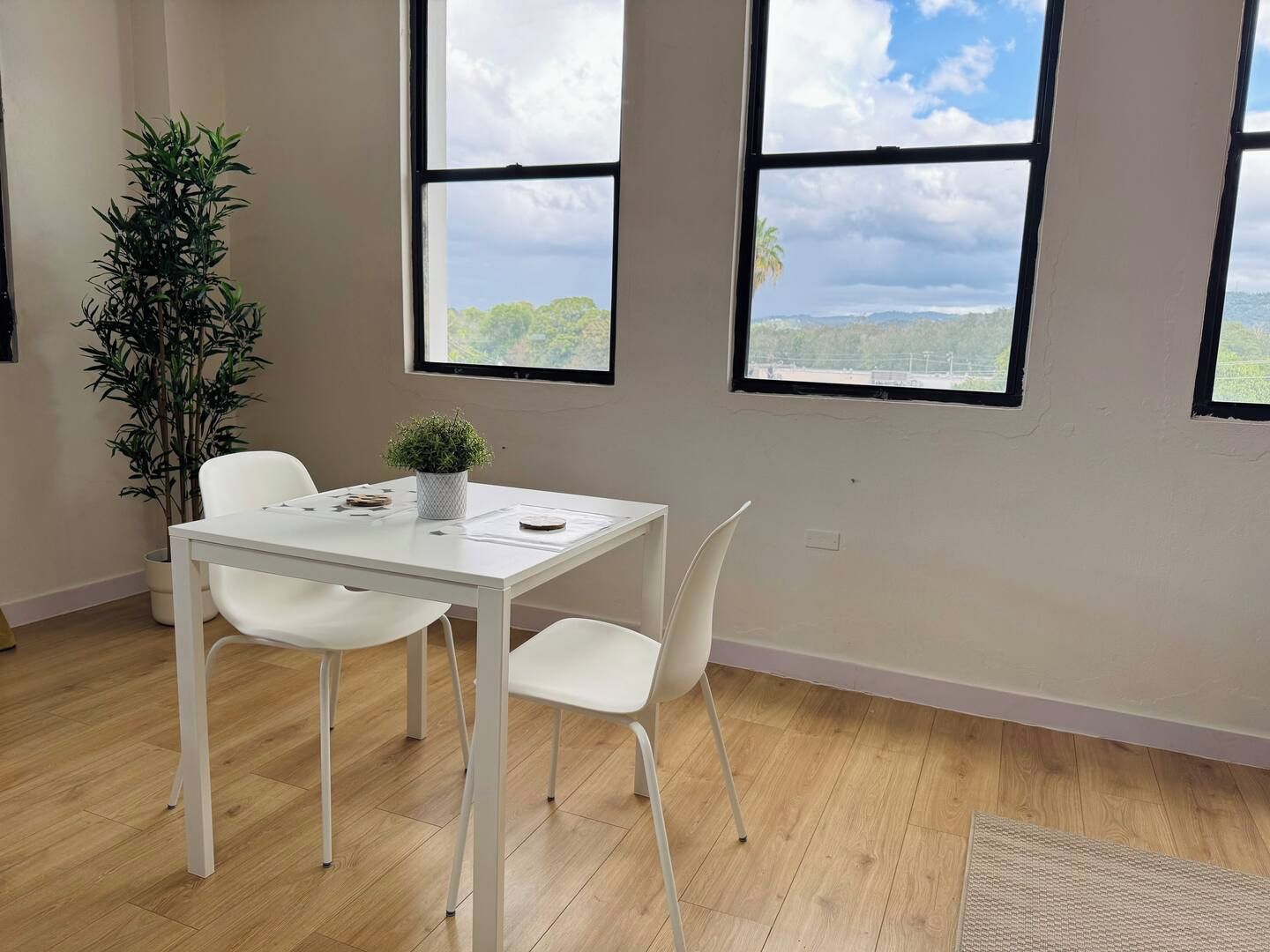 A room with a white table, two white chairs, a potted plant, and large windows with a view of greenery and hills.