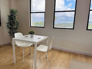 Een kamer met een witte tafel, twee witte stoelen, een potplant en grote ramen met uitzicht op groen en heuvels.