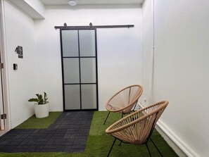 A small, enclosed outdoor area with artificial grass, two wicker chairs, a sliding glass door, and a potted plant.