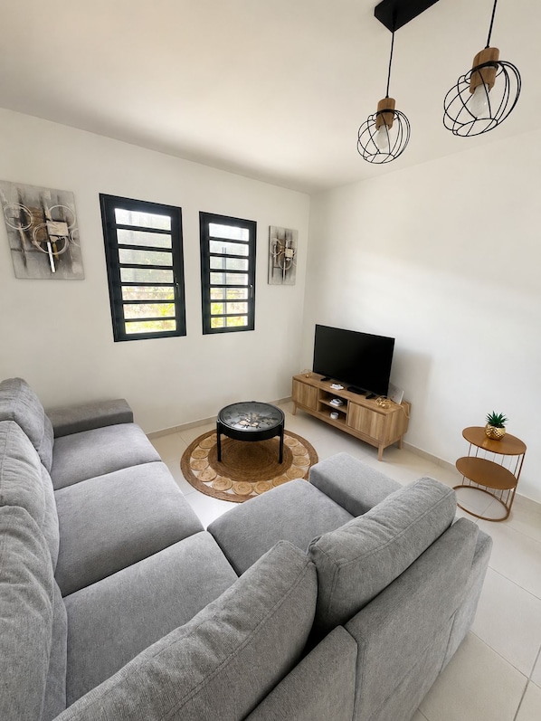 A modern living room with a sectional sofa, a round coffee table, a TV stand, and two windows with blinds. - Le Cocon du Palmier Bleu - Private Pool (Rivière-Salée)