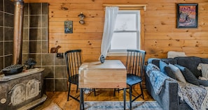 Ein gemütliches Blockhaus-Interieur mit Holzvertäfelung, einem Kamin, einem Esstisch mit Stühlen und einer Couch mit gemustertem Überwurf.