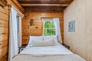 Ein Schlafzimmer einer Holzhütte mit einem Bett, einem Nachttisch, einem Fenster mit Vorhängen und einer Wandleuchte.