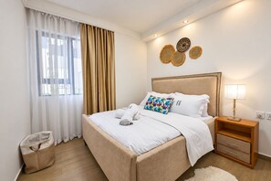 Ein Hotelzimmer mit Bett, Nachttisch, Lampe, Mülleimer und einem Fenster mit Vorhängen.