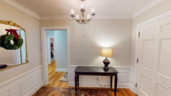 Un couloir orné d’un lustre, une table avec une lampe et un miroir décoré d’une guirlande.