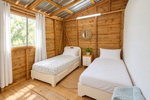 Un dormitorio con dos camas, paredes de madera, techo con vigas de metal, una mesita de noche con una planta y una ventana con cortinas.