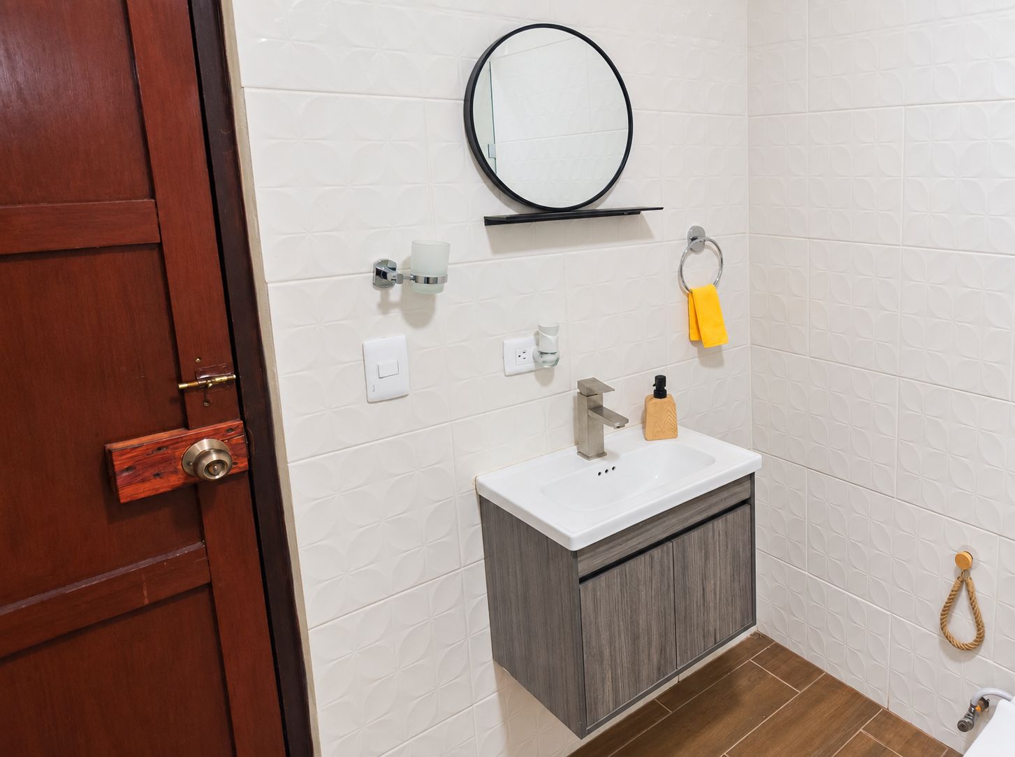 A bathroom with a wooden door, a white tiled wall, a round mirror, a white sink with a wooden cabinet, a towel ring, and a towel.