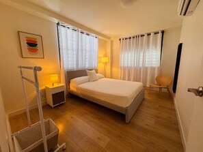 A hotel room with a bed, a chair, a side table with a lamp, a wall-mounted air conditioner, and a painting. - Central San Germán - Private Patio / Generator (San Germán)