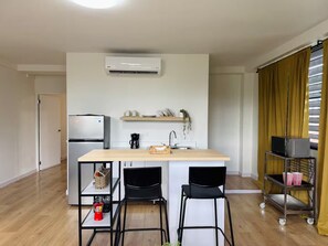 A modern kitchen with a built-in refrigerator, a coffee machine, and a microwave on a shelf. There are two black chairs and a small table in front of the counter. - Central Apt in Historic Town San German - King Bed (San Germán)