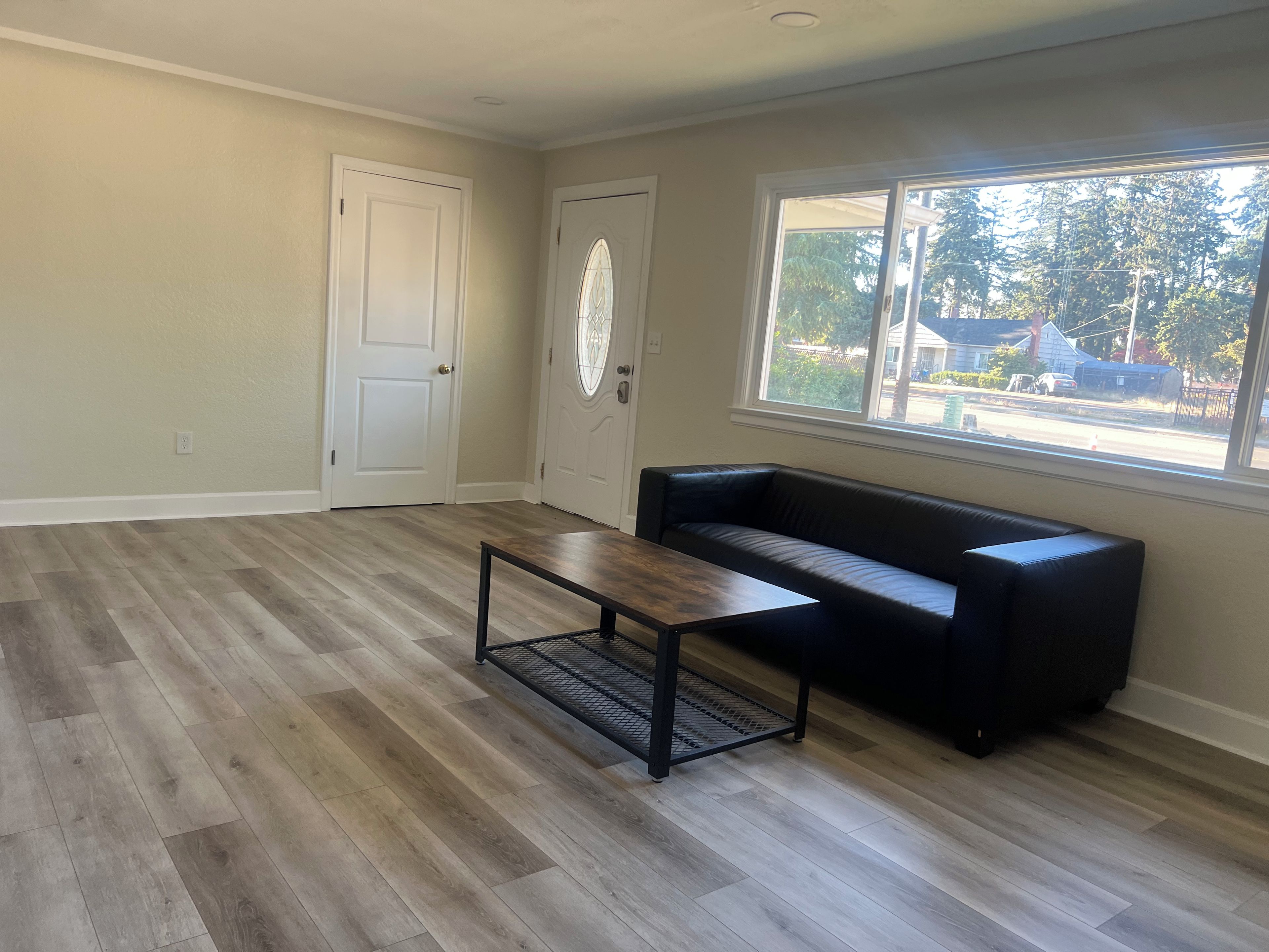 A room with a wooden floor, a black leather sofa, a wooden coffee table, and a window with a view of a residential area.