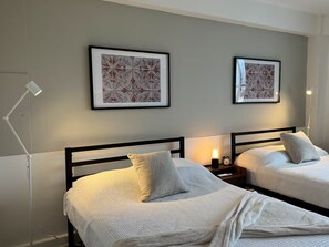 Quarto de hotel com duas camas, cada uma com travesseiro e mesa de cabeceira com luminária.
