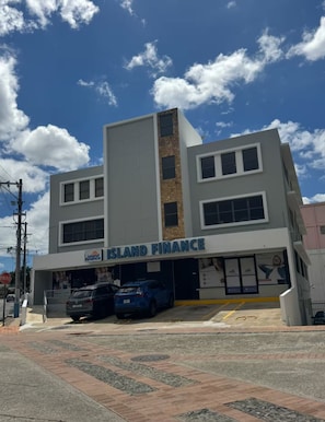 A two-story building with a sign reading 'Island Finance' on the ground floor. There are two cars parked in front. - Historic Stay in San Germán – Modern & Central (San Germán)