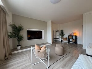 Ein modernes Wohnzimmer mit einem Flachbildfernseher, einem weißen Sessel mit Kissen, einer Topfpflanze und einem Beistelltisch mit Lampe.