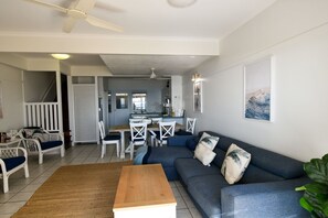 Ein Wohnzimmer mit einer Couch, einem Couchtisch und Sesseln, ein Essbereich mit Tisch und Stühlen sowie eine Küche im Hintergrund.