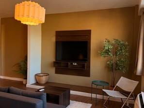 A modern living room with a flat-screen TV mounted on the wall, a dark wooden coffee table, a grey sofa, a white chair, a small round table, and a large potted plant. - Historic San Germán Retreat + Prime Location (San Germán)