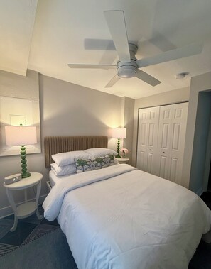 A hotel room with a bed, a ceiling fan, a nightstand with a lamp, a wall-mounted mirror, and a door. - NEW! Brickhouse Villas. 2 blocks from UF. Early check in/late check out. Parking (gainesville)