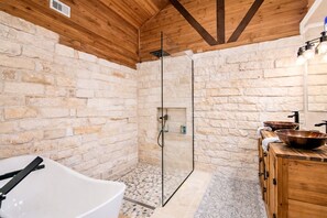 A bathroom with a stone wall, a glass shower enclosure, a wooden vanity with copper sinks, and a white bathtub. - The Limestone Retreat at Ruby Ranch Getaways (Buda)
