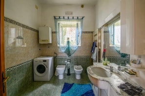 Een badkamer met een wasmachine, twee toiletten, een wastafel en een raam met gordijn.