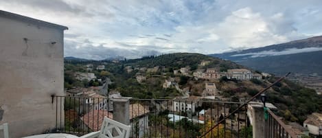 A balcony with a view of a hillside village, a table, and a chair.