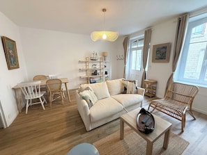 Ein Wohnzimmer mit einer Couch, einem Esstisch mit Stühlen, einem Bücherregal und einem Fenster mit Vorhängen.