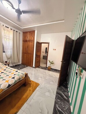 Ein Schlafzimmer mit Bett, Deckenventilator, Fernseher und einem durchgehenden Tür sichtbaren Badezimmer.