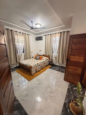 Une chambre à coucher avec un lit, des rideaux, un ventilateur de plafond, un appareil de climatisation et une porte en bois.
