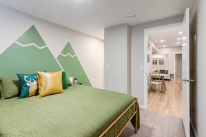 Een slaapkamer met een bed, kussens, een groene wand met een bergdecor en uitzicht op een eetruimte.